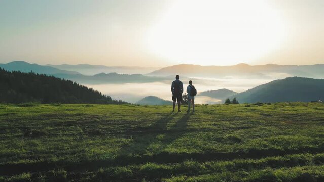 Two tourists on the top of the mountain watching the sunrise and the amazing landscape in the mountains and the fog lying in the valley between the mountains. Nature and beauty of a morning tourist