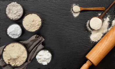 Black bowls with different types of flour, spoon and rolling pin on a black slate background, top view. Space for text.
