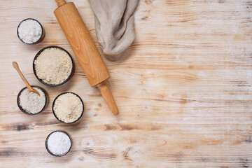 Black bowls with different types of flour, rolling pin and napkin on a white and brown wooden table, top view. Space for text.