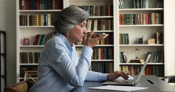 Serious older grey haired business woman talking on mobile phone on speaker, typing on laptop computer at office workplace, recording audio message, making call. Elder professional concept