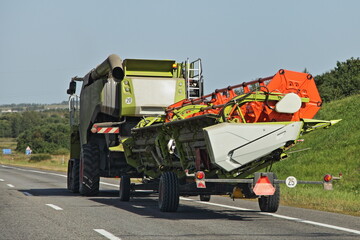 Modern European Harvester combine drive with reaping machine on trailer on countryside asphalt road at summer day - harvesting, agriculture, farming in East Europe