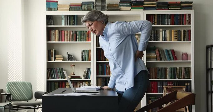 Exhausted unhappy senior business lady feeling strong back ache after sitting at workplace, touching lumbar massaging spasm muscles, suffering from pain attack at laptop in office