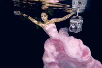 young beautiful woman in long  pink dress with cage in her hands underwater