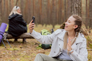 Satisfied woman with blonde hair tied in braids sits on the ground in the forest talking on the...