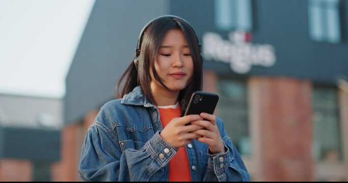 Portrait of Asian girl teenager in headphones, using smartphone while walking