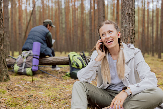 Woman with blonde hair tied in braids sits on the ground in the forest and talks on the phone, resting while trekking, camping, hiking.