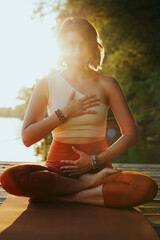 A calm woman in the lotus pose sits on a dock and practices yoga breathing near the river.