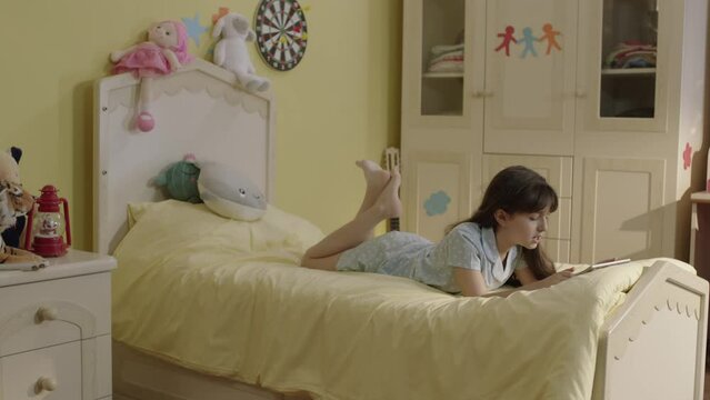 Little girl surfing internet with tablet computer lying on her bed at home.Cute girl continues distance education on tablet in bedroom. She watches funny videos and plays online games she.