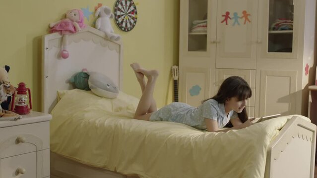 Little girl surfing internet with tablet computer lying on her bed at home.Cute girl continues distance education on tablet in bedroom. She watches funny videos and plays online games she.