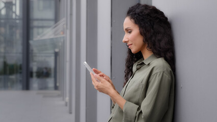 Latin Caucasian Hispanic girl woman 30s businesswoman holding cell phone standing near wall building outdoors success good news mobile app win surprised lady celebrating victory on smartphone triumph