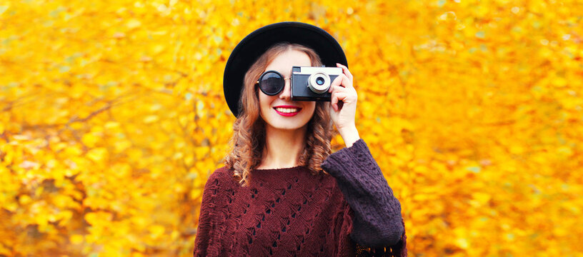 Autumn portrait of happy smiling young woman photographer with film camera wearing round hat, knitted poncho in the park on yellow leaves background
