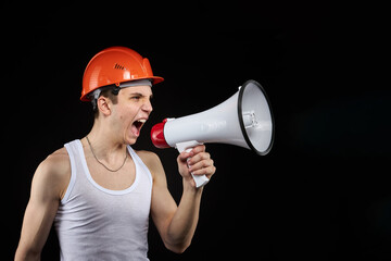 Fototapeta premium A teenager in an orange construction helmet with a loudspeaker in his hand on a dark background shouts into a megaphone