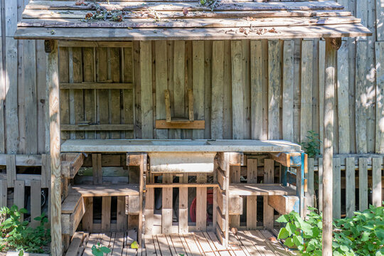 Wooden booth in neat and clean work area of a vegetable plot against wall of wooden planks, withe table, sink, sloping ceiling with remains of fallen branches, sunny summer day in an organic orchard