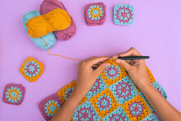 Top view of hands crocheting multicolored granny squares afghan on light violet background with cotton balls and small crochet pieces around.