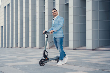 Joyful businessman with scooter dressed in blue suit and sunglasses against city building.