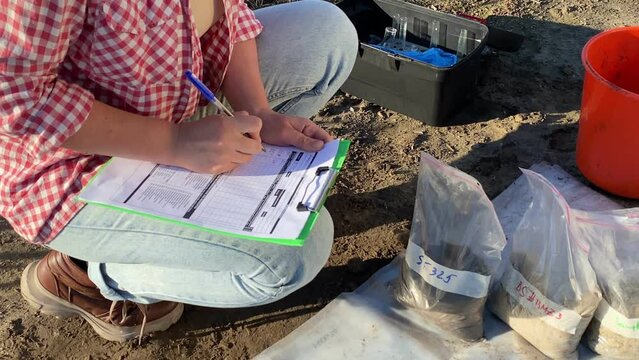 Closeup of female agronomist preparing soil samples for laboratory analysis, writing in information sheet outdoors at sunrise. Woman farmer taking notes in form, working at field. Soil certification