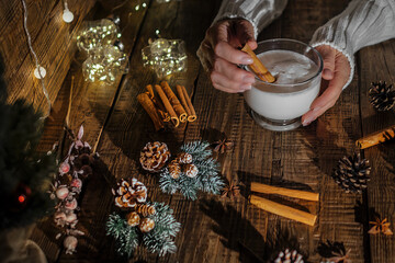 Female hands in a white sweater hold a glass cup in which a festive Christmas hot drink eggnog is poured on a wooden background with fir branches and cinnamon