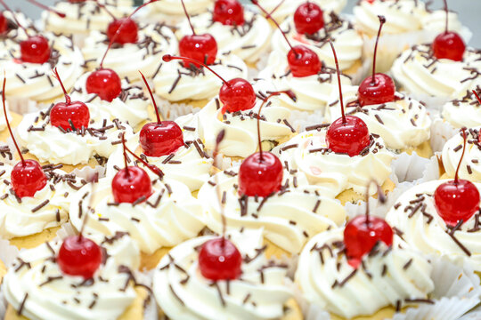 Various mini cakes with whipped cream, chocolate sprinkles and cherries on top, as the focus on these.