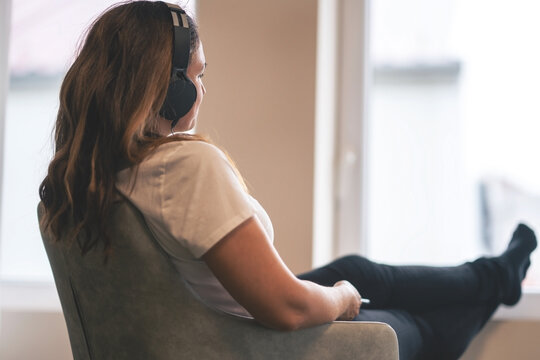 She listens to music on headphones and looks thoughtfully out the window.A woman is sitting on a chair listening to music and looking into the distance through the window. - Powered by Adobe