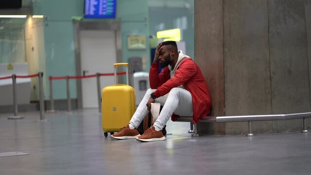 Sad African American man upset at airport his flight is delayed. Depressed traveler male waiting for a plane sitting in empty terminal with baggage. Exhausted guy on a long night connection at airport
