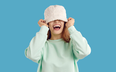 Happy funny carefree joyful young woman or teen girl with toothy smile puts on white cozy warm fluffy fuzzy artificial teddy lamb wool bucket hat that covers her eyes. Fashion concept Studio head shot - obrazy, fototapety, plakaty