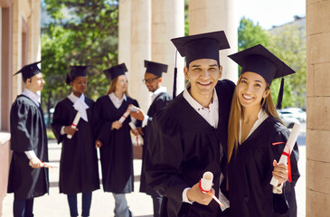 Happy friends on graduation day. Portrait of two cheerful joyful students standing near university building with other graduates in background, holding diplomas, hugging, smiling and looking at camera