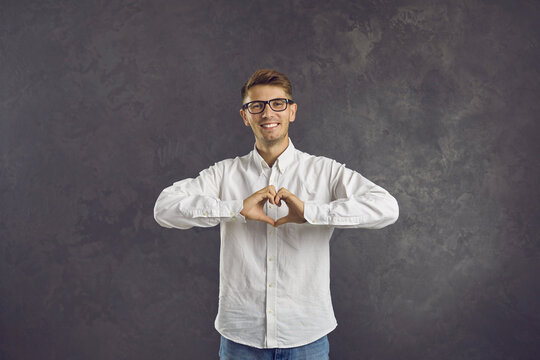 Thank You, I Really Appreciate It. Happy Man Forming Heart Shape With Hands Standing On Grey Background. Positive Guy In White Shirt And Glasses Doing Heart Sign To Show Love, Support And Kindness