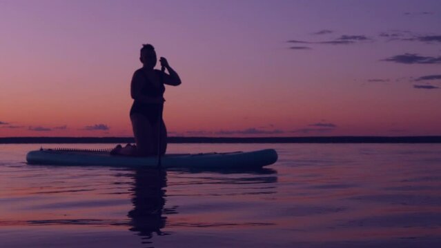 A woman in a bathing suit with a Mohawk floats on a SUP board on the lake at sunset against a purple sky. Overall plan