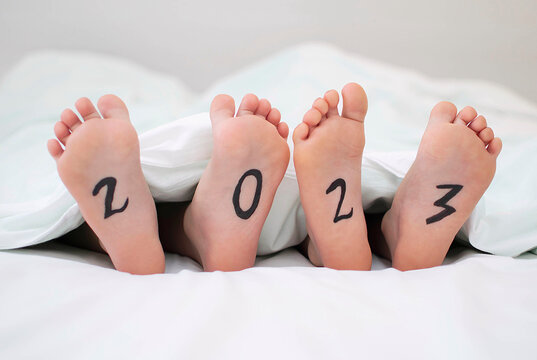 Close-up of the bare feet of two children sticking out from under a white blanket on a bed with the inscription 2023. the concept of the winter New Year. space for text