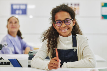 Portrait of smiling black schoolgirl wearing glasses sitting at desk in classroom and looking at camera