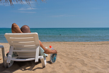 A man lies on a white sun lounger on a sandy beach near the sea under a straw umbrella. A snorkeling mask with protection against waves and fog hangs on a sun lounger.