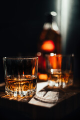 A glass of whiskey on a wooden table. In a glass filled with alcohol from the bottle. In the background, two bottles of whiskey of different shapes, a glass of whiskey and empty glasses of whiskey.