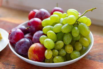 green grapes and plums in a plate on a wooden board, windowsill, concept of fresh fruits and healthy food