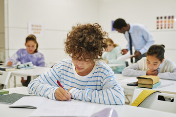 Portrait of young schoolboy with curly hair sitting at desk in school classroom and writing in notebook