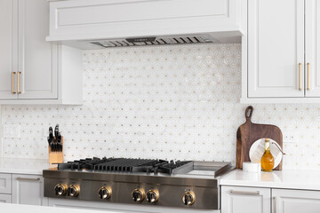 A detail shot of a beautiful kitchen's stainless steel luxury stove, hood, granite counter tops, and a custom, white and gold tiled back splash.