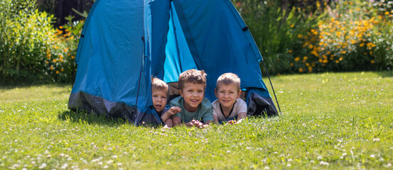 Three little boys lie in a blue tourist tent in the yard.