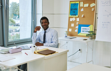 Portrait of young black teacher smiling at camera while sitting at desk in empty classroom, copy space