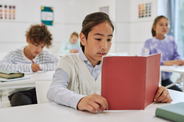 Portrait of teenage boy sitting at desk in school classroom and reading book with group of kids in background
