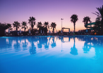 Fototapeta na wymiar Beautiful reflection in swimming pool at colorful sunset. Purple sky reflected in water, palm trees, sun beds, umbrellas at night in summer. Luxury resort. Landscape with empty pool in twilight