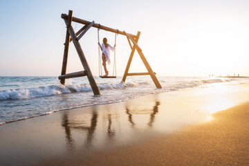 Happy young woman on wooden swing in water, beautiful blue sea with waves, sandy beach, reflection in water, golden sky at sunset. Summer in Side, Turkey. Girl ride on a swing on sea coast. Travel