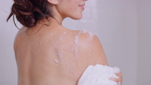 Beautiful brunette woman washes shoulder in shower with body wash and lufa