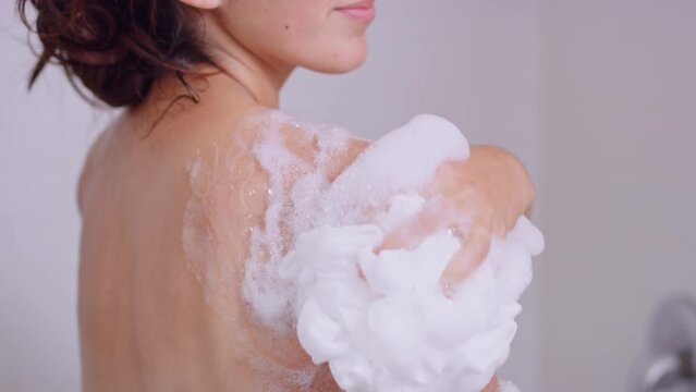 Close on woman's shoulder in shower as she slowly scrubs body wash bubbles