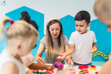Obraz na płótnie Canvas Relaxing sensory play with moldable kinetic sand at nursery school. Toddlers with their teacher having fun around the table using different tools for sculpting sand such as colorful and textured