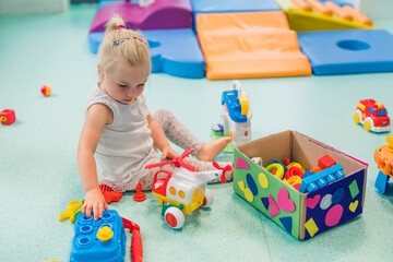 Fototapeta premium Toddler girl playing with different plastic toys such as building blocks, car toys. Work on problem-solving skills, shape and color recognition, fine motor and gross motor skills development through