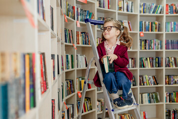 girl with glasses reads a book sitting on a stepladder in the library