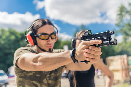 Caucasian bearded man in safety gear and camouflage clothing aiming handgun. Firearms training at outdoor shooting range. Horizontal shot. High quality photo