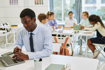 Portrait of young black teacher using laptop in classroom with group of children in background