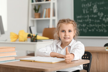 Cute little first-grader showing thumb-up at desk in classroom