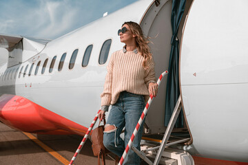 Woman passenger in sunglasses standing on airplane stairs at airport and looking away