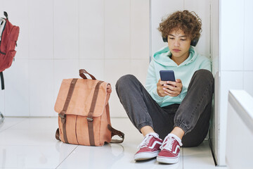 Full length portrait of isolated teen schoolboy sitting alone in corner and using smartphone, copy...